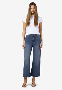 Γυναίκα όρθια που φορεί ένα λευκό κοντομάνικο μπλουζάκι, κοντές φαρδιές τζιν παντελόνες σε μπλε χρώμα με ακατέργαστα τελειώματα και καφέ σαγιονάρες, μπροστά σε λευκό φόντο.