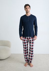 Man staat blootsvoets in een kamer met een lichttapijt en een stoel, gekleed in een marineblauw langshirt en rood, wit en blauw geruite pyjamabroek.