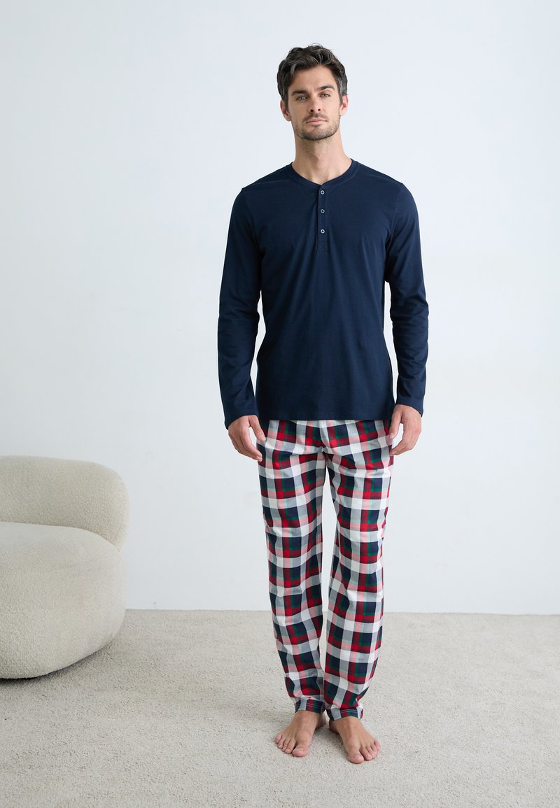 Man staat blootsvoets in een kamer met een lichttapijt en een stoel, gekleed in een marineblauw langshirt en rood, wit en blauw geruite pyjamabroek.