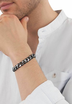 Hombre con camisa blanca y pulsera de cadena negra y plateada en la muñeca, con la mano apoyada en el mentón en una pose pensativa.