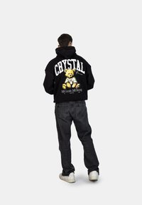 Felpa nera oversize con una grafica di un orsetto e il testo "CRYSTAL" sul retro. Indossata con jeans scuri e sneakers bianche.