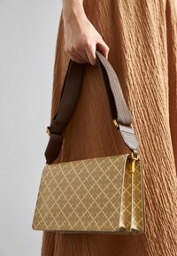 Beige kankuri kaboji yenye muundo wa kijiografia, ikiwa na mkanda wa kahawia wenye texture na mapambo ya dhahabu. Mukono wa kubeba unaonekana.