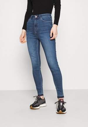 Osoba stojící před bílým pozadím, oblečená v modrých džínech s vysokým pasem a černo-šedých teniskách New Balance.