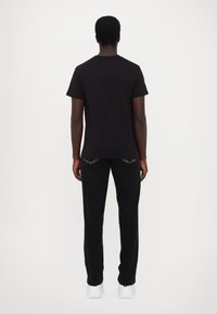Tricou din bumbac negru cu mâneci scurte, asociat cu pantaloni negri. Ținuta este completată cu teniși albi, văzuți din spate.
