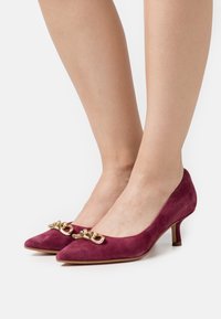 Escarpins en suede bourgogne avec un bout pointu, un talon bas et un accent en chaîne dorée sur le devant. Texture lisse et design épuré.