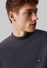 Maglione in pile blu navy con colletto alto e superficie testurizzata. Presenta un piccolo logo black di Calvin Klein sul lato sinistro del petto.