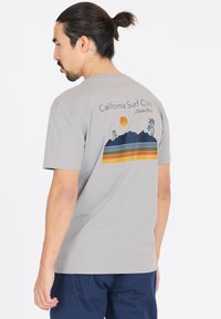 Harmaa puuvillainen t-paita lyhyillä hihoilla, jossa on selässä monivärinen graafinen kuvasto vuorista, palmupuista ja auringosta.