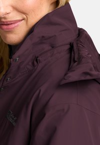 Violette wasserdichte Jacke mit hohem Kragen, Druckknöpfen und einer Reißverschlusstasche. Mit Logo-Stickerei auf der Brust und glatter Textur.