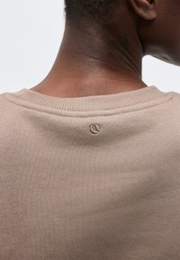 Κοντινό πλάνο του λαιμού και του άνω μέρους της πλάτης ενός ατόμου που φοράει μπεζ πλεκτό πουλόβερ με ένα μικρό κεντημένο λογότυπο κάτω από το γιακά.