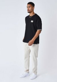 T-shirt nera con maniche corte, bordi bianchi sui polsini e una piccola toppa con logo; abbinata a pantaloni beige chiaro e sneakers bianche.