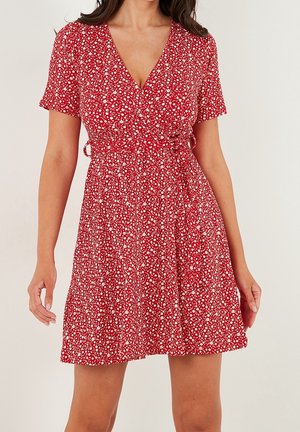 Nainen, joka käyttää punaista, lyhythihaista v-kaula-aukkoinen mekkoa, jossa on valkoinen kukkakuvio ja vyötäröllä sidottava nauha.