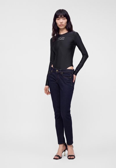 Femme debout portant un body noir à manches longues avec un texte blanc et un jean bleu foncé, associé à des sandales noires à talons hauts.