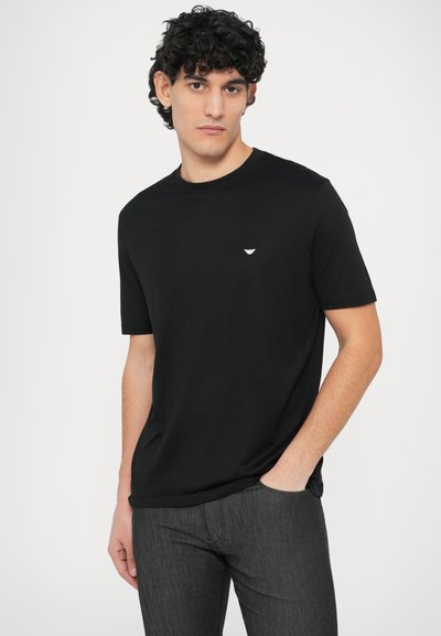 Črna bombažna majica s kratkimi rokavi in okroglim izrezom. Na prsih ima majhen bel logotip. Kombinirana s temno sivimi teksturiranimi hlačami.