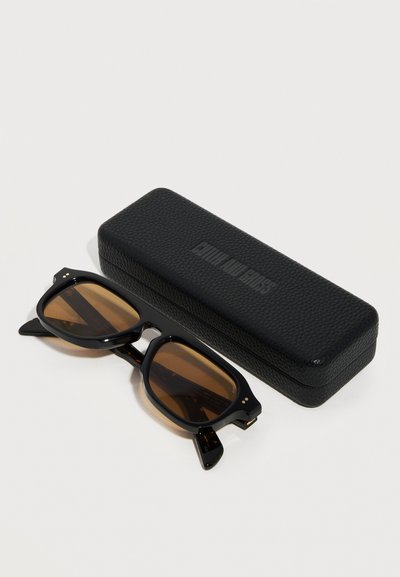 Occhiali da sole rettangolari neri con lenti marroni appoggiati accanto a una custodia rigida nera texturizzata su uno sfondo grigio.