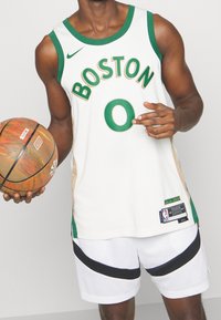 Maglia da basket bianca con accenti verdi e testo "BOSTON", abbinata a pantaloncini bianchi in rete con una striscia nera. Tiene in mano un pallone da basket multicolore.