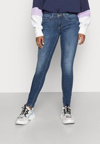 Jeans skinny em denim azul escuro, com cinco bolsos, cintura média e ligeira descoloração. Combinados com sapatilhas robustas.