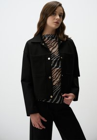 Zwarte cropped jack met gouden drukknoopjes, gecombineerd met een patroon top met strepen en spikkels in bruin- en zwarttonen.