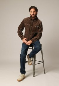 Bruine corduroy shirtjack met twee voorzakken, knoopsluiting en een gestructureerde afwerking. Draag met blauwe jeans en beige sneakers.