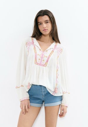 Jeune femme aux longs cheveux bruns portant un chemisier blanc brodé et ample, ainsi qu'un short en denim bleu retroussé, debout devant un fond blanc.