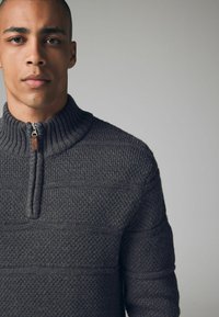 Maglione grigio scuro a maglia con colletto alto a coste, chiusura a zip quartocento e motivo testurizzato, che mette in mostra diversi disegni di punto.