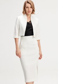 Veste blanche cropped avec un large col, associée à une jupe midi blanche ajustée avec une fente sur le côté. Débardeur en dentelle noire en dessous.