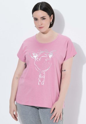 Frau mit kurzen dunklen Haaren, die ein pinkes T-Shirt mit einer weißen Giraffenskizze und graue Hose trägt, steht vor einer weißen Wand.