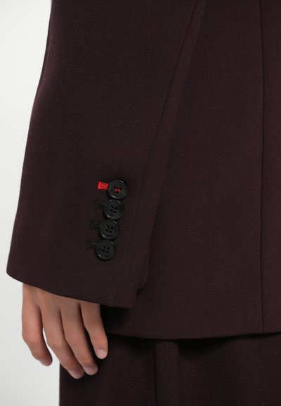 Rukávy burgundské saka mají čtyři černé knoflíky, dekorativní šití a malý červený akcent blízko manžety. Hladká textilie.