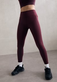 Bordeaux leggings med høy midje, glatt tekstur og et tettsittende design, parret med svarte treningssko og hvite sokker.