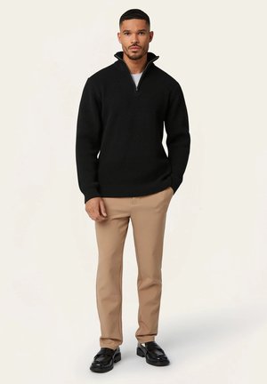 Black ribbed knit pullover sweater with a high collar and zipper, paired with beige pants and black loafers, standing against a light background.