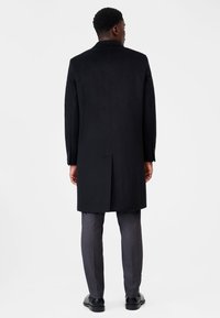 Svart överrock med en strukturerad design, som har rak passform, långa ärmar och en slits bak, tillsammans med grå byxor.