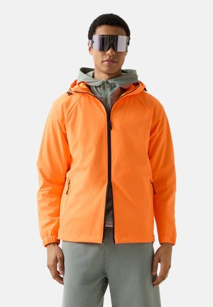 Jonge man draagt een feloranje jas met rits, een grijze capuchontrui, grijze broek en grote reflecterende zonnebril, staand tegen een effen achtergrond.