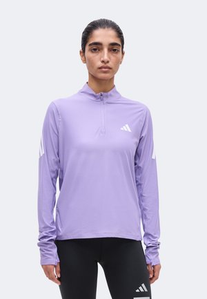 Femme portant un top de sport lavande à manches longues avec une fermeture éclair au quart et un legging noir, debout devant un fond clair uni.