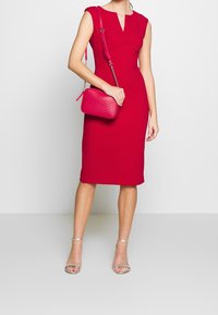 Robe rouge ajustée avec des manches courtes et un décolleté en V ; associée à un sac bandoulière matelassé rose et des sandales argentées à lanières.