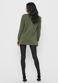 Top de mangas longas verde oliva com um corte descontraído, apresentando um decote redondo e detalhe na costura das costas, combinado com leggings pretas justas e saltos altos.