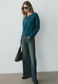 Cardigan turchese con bottoni, abbinato a jeans a gamba larga e sbiaditi. La modella tiene una borsa tote nera; lo sfondo è grigio chiaro.