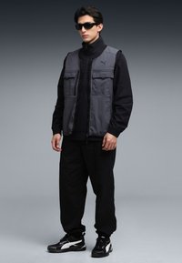 Grijze textielen vest over een zwart topje met rits, gecombineerd met zwarte broek en zwarte sneakers. Voorzien van meerdere zakken aan de voorkant en een hoge kraag.