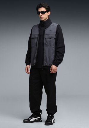 Szara tekstylna kamizelka na czarnej bluzie na zamek, w połączeniu z czarnymi spodniami i czarnymi trampkami. Posiada wiele przednich kieszeni oraz wysoki kołnierz.