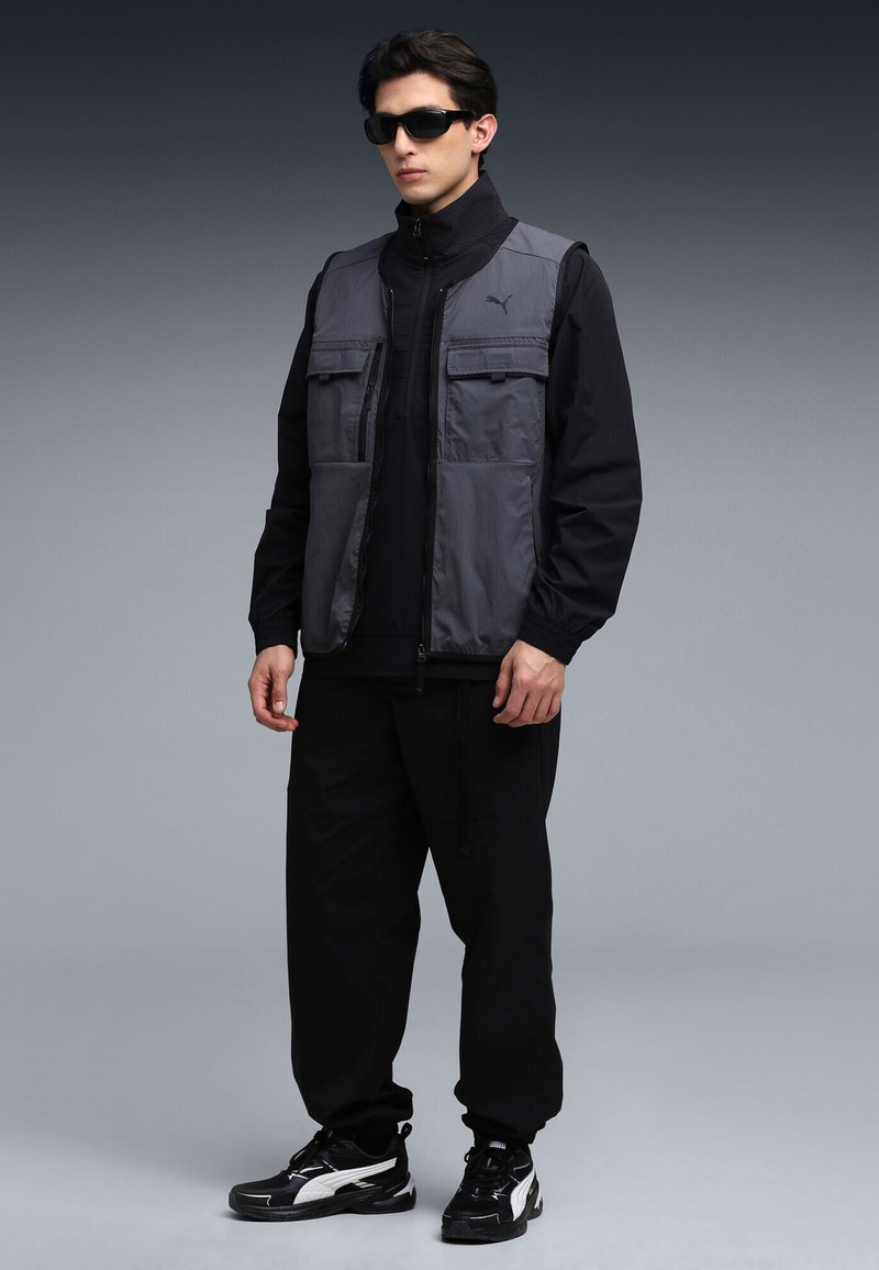 Grijze textielen vest over een zwart topje met rits, gecombineerd met zwarte broek en zwarte sneakers. Voorzien van meerdere zakken aan de voorkant en een hoge kraag.