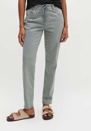 Osoba nosząca jasnoszare spodnie z troczkami i beżowe sandały z odkrytymi palcami i złotymi klamrami, stojąca na jednolitym tle.