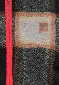 Fleece bunda s čierno-šedo-hnedým károvaným vzorom, so červeným zipsom a hnedou koženou nášivkou na hrudi.