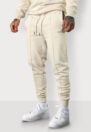 Beige Jogginghosen aus strukturiertem Stoff, mit seitlichen Taschen, schmal zulaufendem Schnitt und dezentem "ESTEEM" Stick. Kombiniert mit weißen Sneakers.