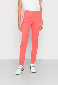 Person som bär korallrosa Adidas träningsbyxor med vita ränder på sidorna och vita Adidas sneakers med svarta ränder.