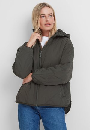 Blonde woman wearing a dark gray quilted jacket with hood, white shirt, and blue jeans, looking to the side with hand near face.
