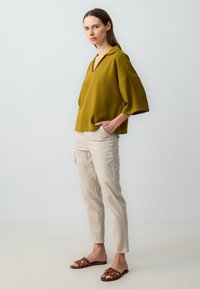 Mosterdgroene, relaxte top met V-hals en brede mouwen, gecombineerd met lichtbeige slim-fit broek en bruine platte sandalen met bandjes.