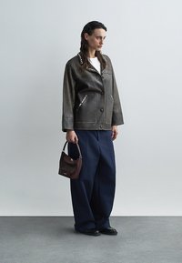 Bruine leren jas met metalen accenten, diepblauwe wijde broek en een kleine bordeauxrode handtas. Model staat op een grijze vloer.