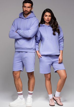 Helle lila Baumwoll-Hoodies und Shorts mit Kängurutaschen. Das männliche Modell trägt weiße Sneaker; das weibliche Modell trägt rosa Sneaker.