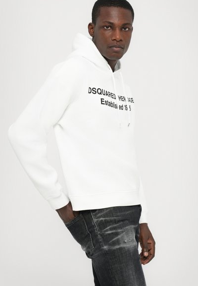 White hoodie with a large black printed logo on the front, paired with dark denim jeans featuring distressed accents and a slim fit.