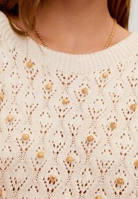 Sweter z dzianiny w kolorze kremowym, z otwartym wzorem w diamenty, ozdobiony beżowymi okrągłymi koralikami, posiadający ściągacz przy okrągłym dekolcie.