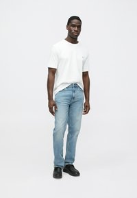 Jaunas vyras stovintis atsipalaidavusiu pozu, dėvintis baltą trumpomis rankovėmis marškinius, šviesiai mėlynas džinsus ir juodus batus, priešais paprastą foną.