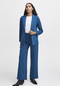 Türkisen Anzug, bestehend aus einem maßgeschneiderten Blazer und weiten Hosen, kombiniert mit einem weißen Oberteil. Hergestellt aus einem glatten, dehnbaren Stoff. Schwarze Knöchelstiefel.